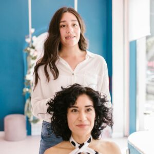 Two women in a hair salon, one styling the other's hair, both smiling brightly.
